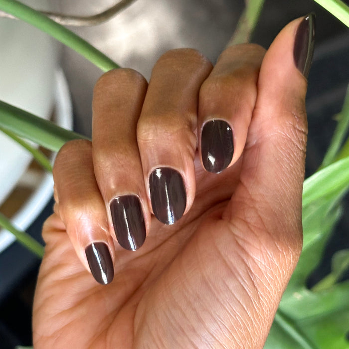 Hand with Dark Chocolate polish on short oval nails against blurred green plant background for rich manicure look