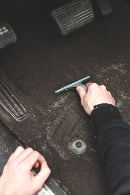 A gloved hand using a Detail Factory Pet Hair Rake on the carpeted ground of a vehicle.