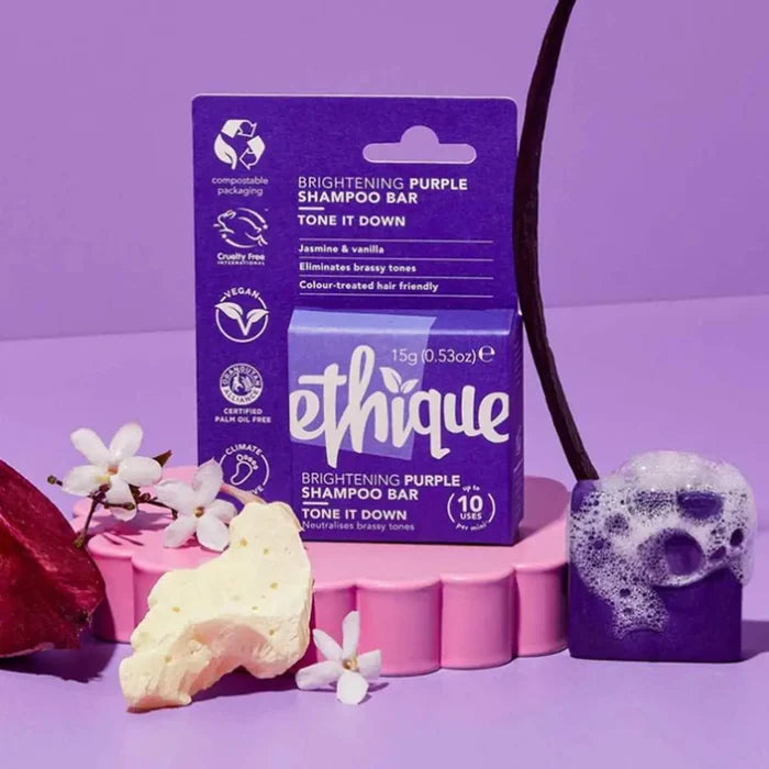 Ethique Brightening Purple Shampoo Bar packaging on a purple background with jasmine flowers, vanilla bean, cocoa butter and foaming bar of purple shampoo.