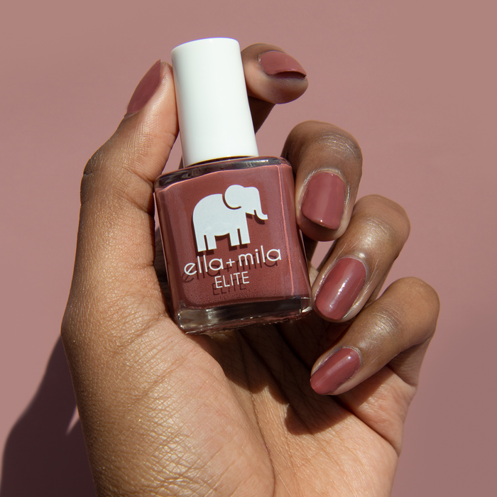 Wanderer brick red nail polish on a deep skin tone, holding the bottle on a mauve background