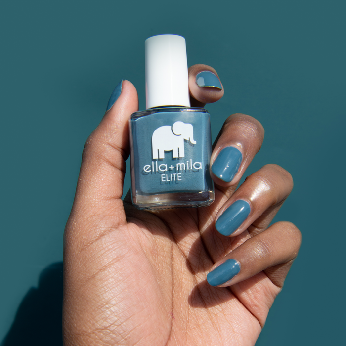 Window Seat grey blue nail polish on a deep skin tone, holding the bottle on a teal background